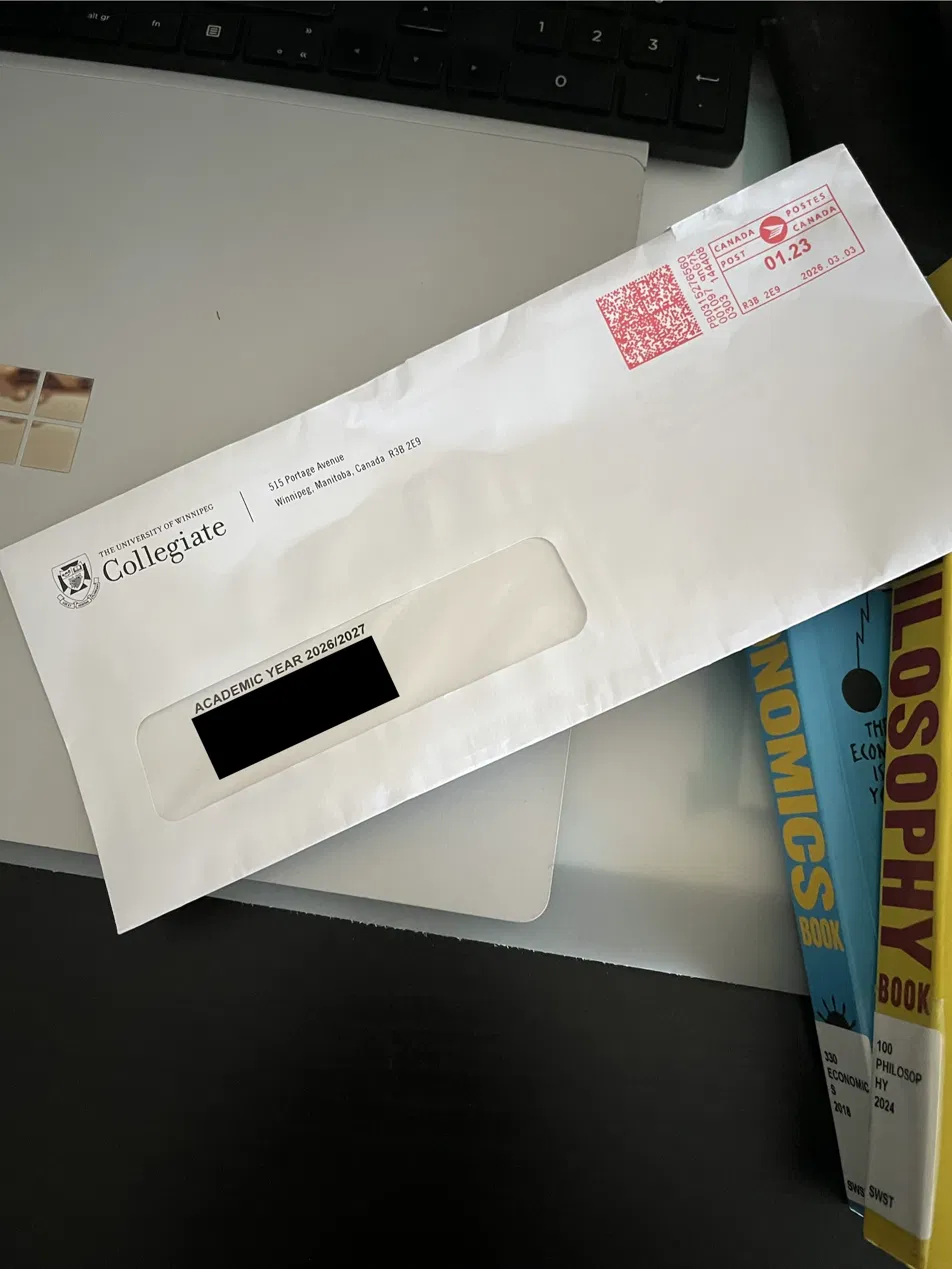 University of Winnipeg Collegiate acceptance envelope resting on a desk with an economics book and a philosophy book visible underneath.